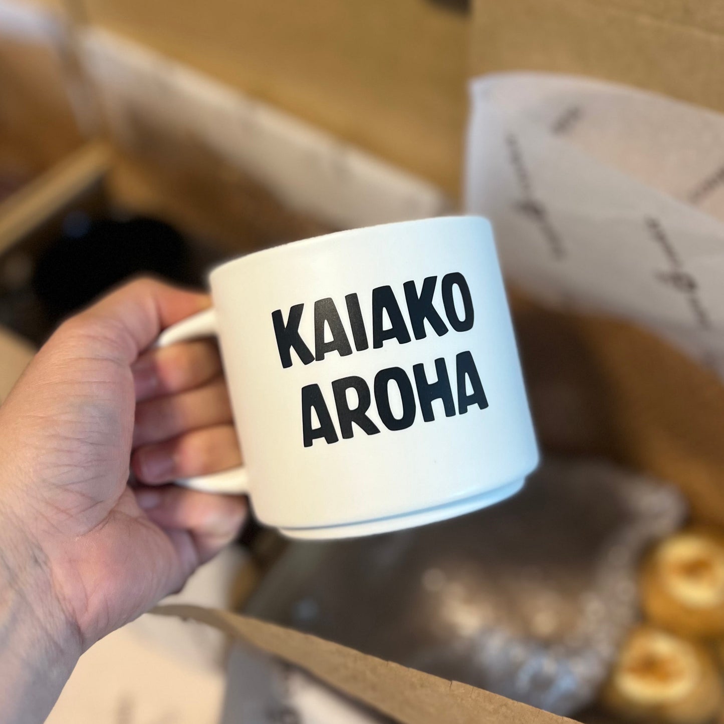 White mug with 'KAIAKO AROHA' text held by a hand, blurred background