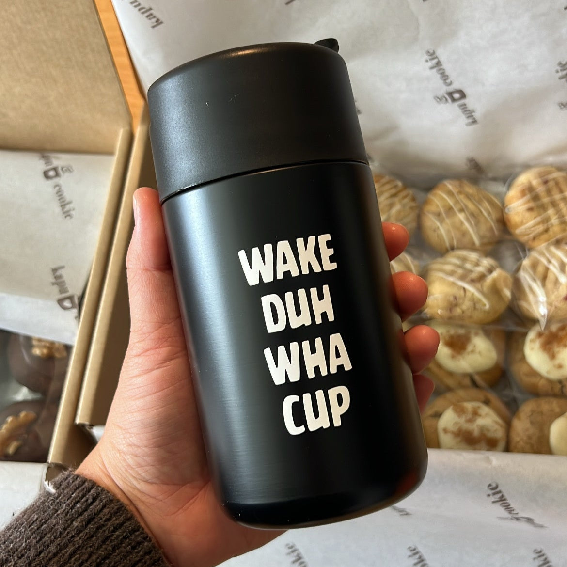 Black thermos with 'Wake Duh Wha Cup' text held in a hand, with cookies in the background.